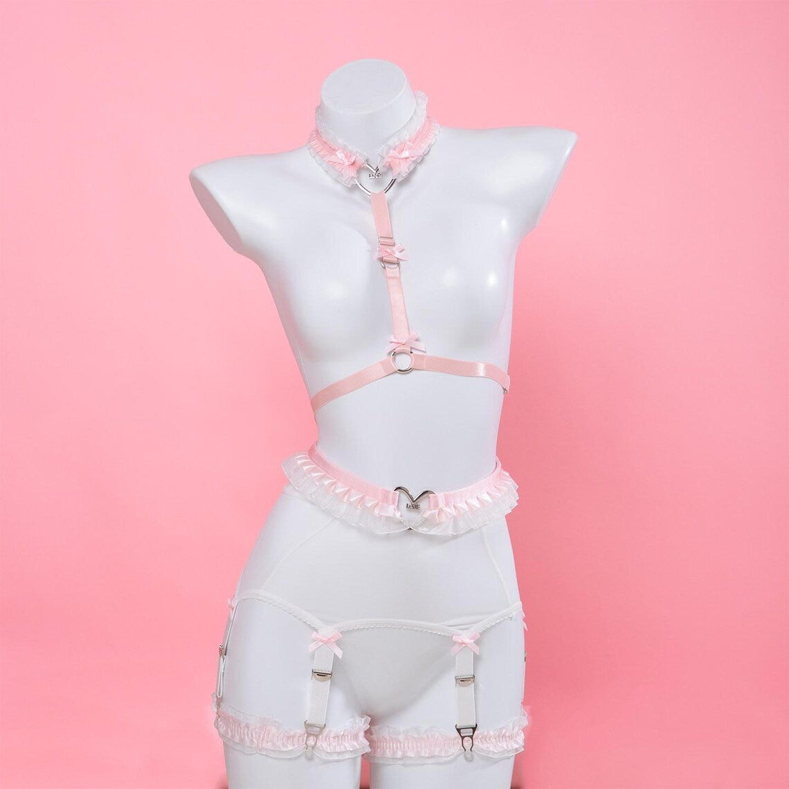 pink and white harness set featuring a ruffled chest harness, heart ring waist belt and white mesh suspender belt, displayed on a white mannequin against a pink background.