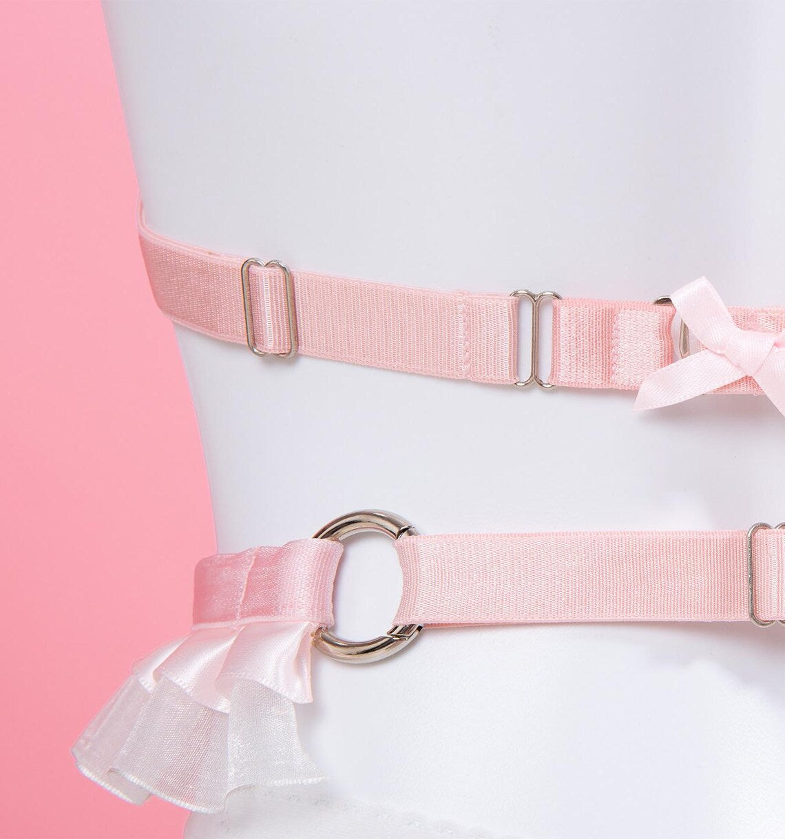 close-up rear view of a pink and white ruffled harness set, showing the silver tone hardware, adjustable sliding straps and pink satin bow applique, displayed on a white mannequin against a pink background.