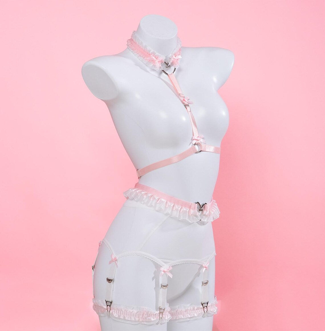 pink and white harness set featuring a ruffled chest harness, heart ring waist belt and white mesh suspender belt, displayed on a white mannequin against a pink background.
