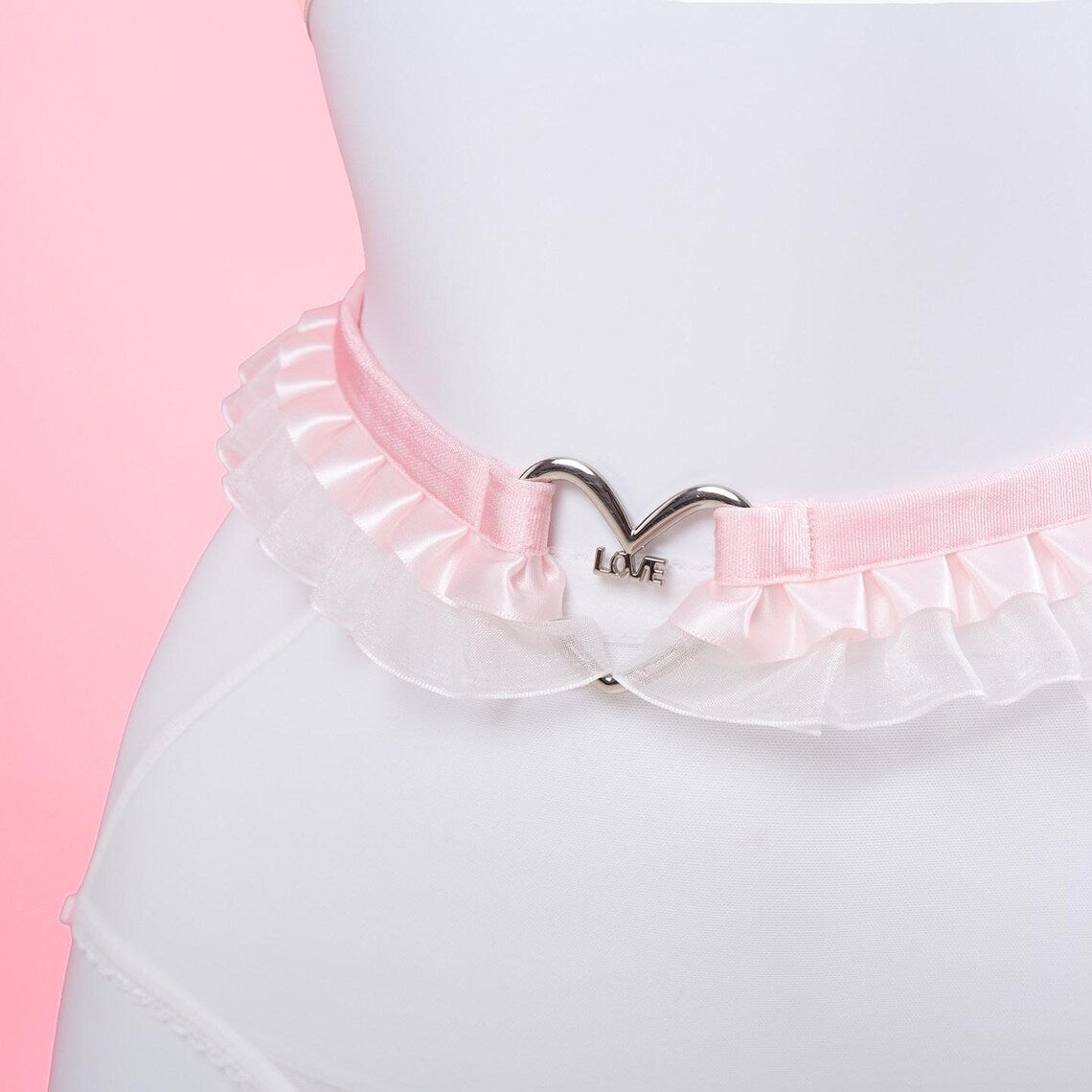 close-up view of a pink and white ruffled waist belt as part of an elasticated harness set, showing the central silver heart ring and ruffled lace trim, displayed on a white mannequin against a pink background.
