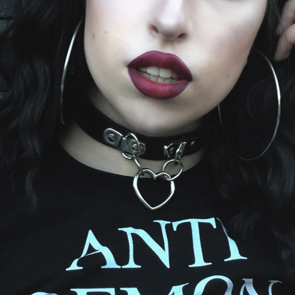 Modelled photo of a black leather collar featuring a central silver heart ring suspended by two D ring plates. Styled with a black graphic t-shirt and dark lipstick for a gothic aesthetic.