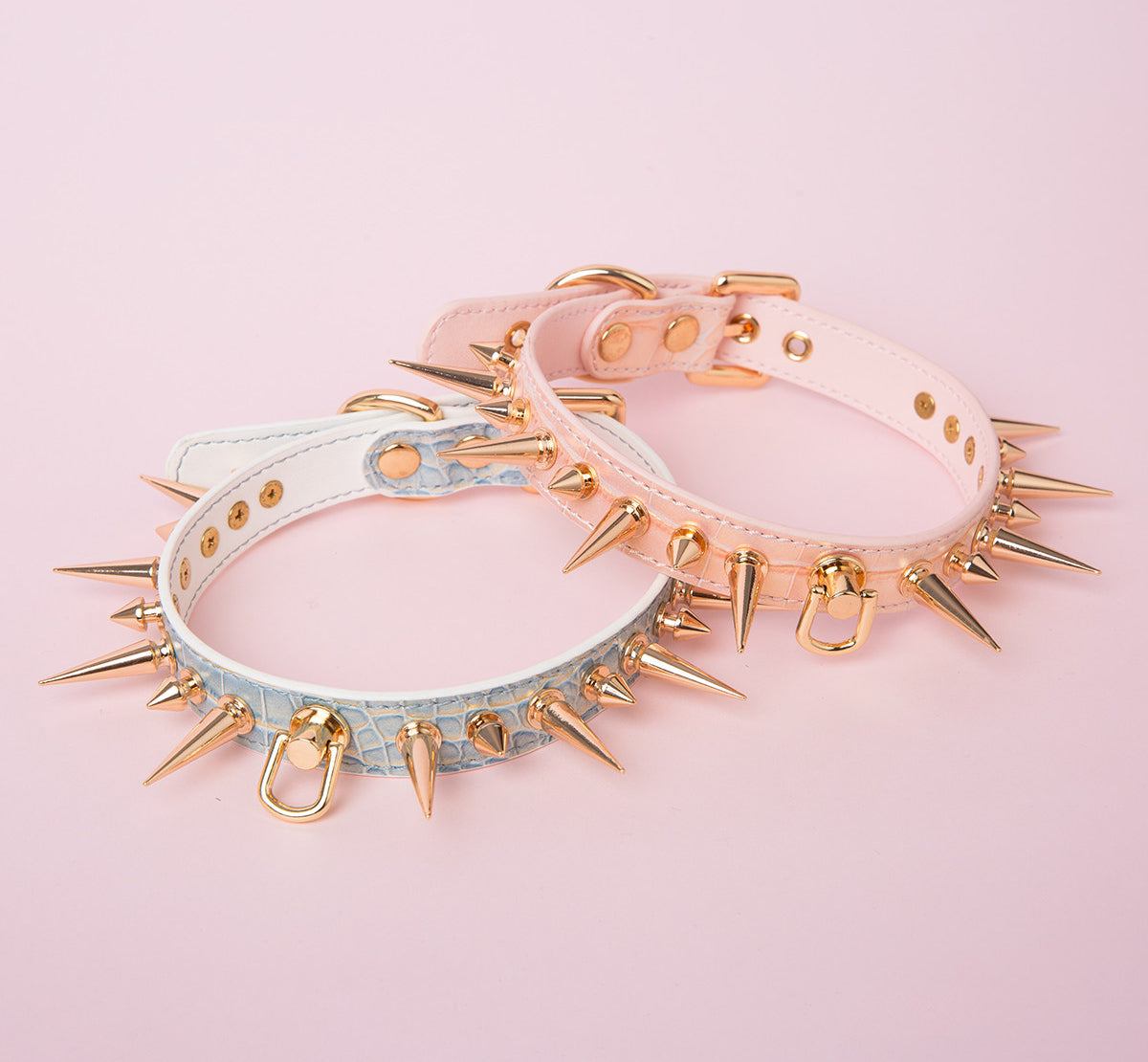 two blue and pink leather collars featuring gold-tone alternating length spike studs and central D ring attachments, displayed on a pink background.