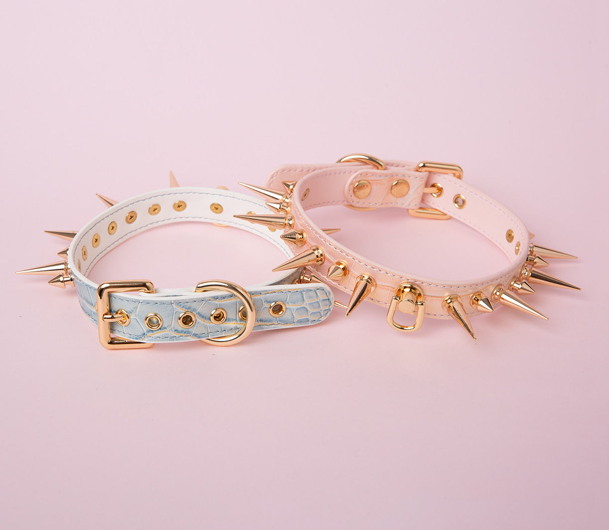 two blue and pink leather collars featuring gold-tone alternating length spike studs, central D ring attachments and adjustable buckles at the back, displayed on a pink background.