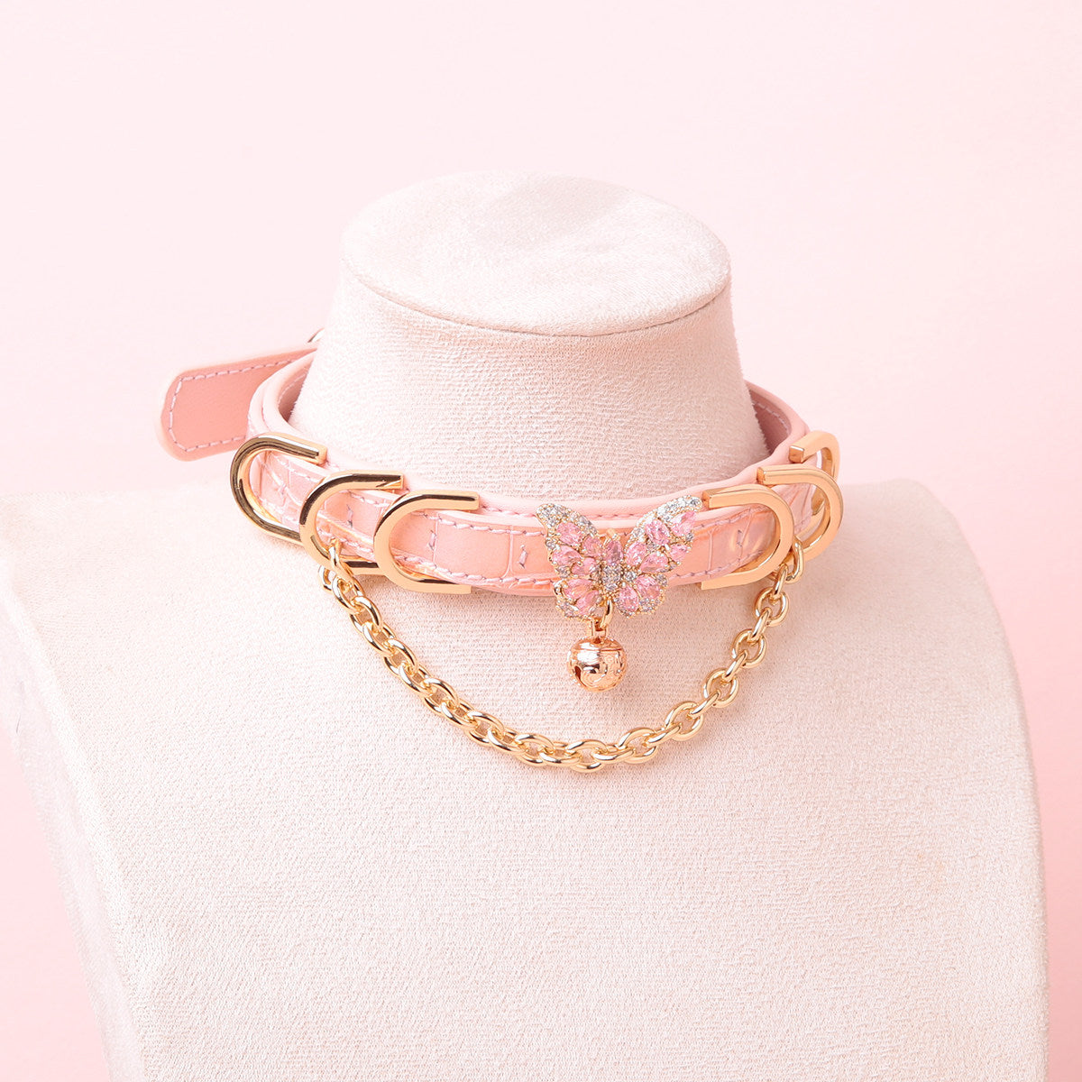 pink leather collar featuring a gold and pink diamante butterfly charm, chain, D rings and bell, displayed on a white mannequin against a pink background.