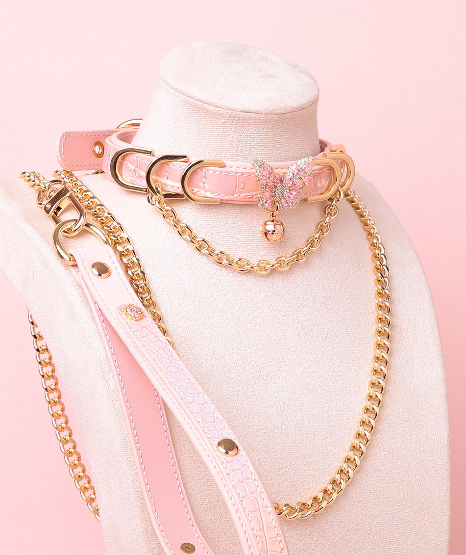 pink and gold leather collar and leash set featuring a gold and pink diamante butterfly charm, gold tone chains, D rings, detachable chain leash and leather hand strap, displayed on a white mannequin against a pink background.
