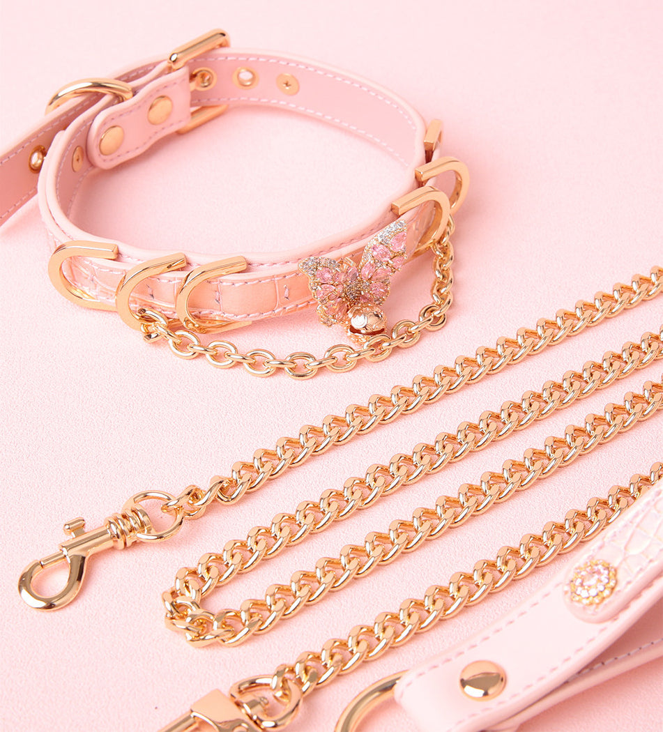 pink and gold leather collar and leash set featuring a crystal butterfly pendant, gold chain, D rings, bell, detachable chain leash and leather hand strap, displayed on a pink background.
