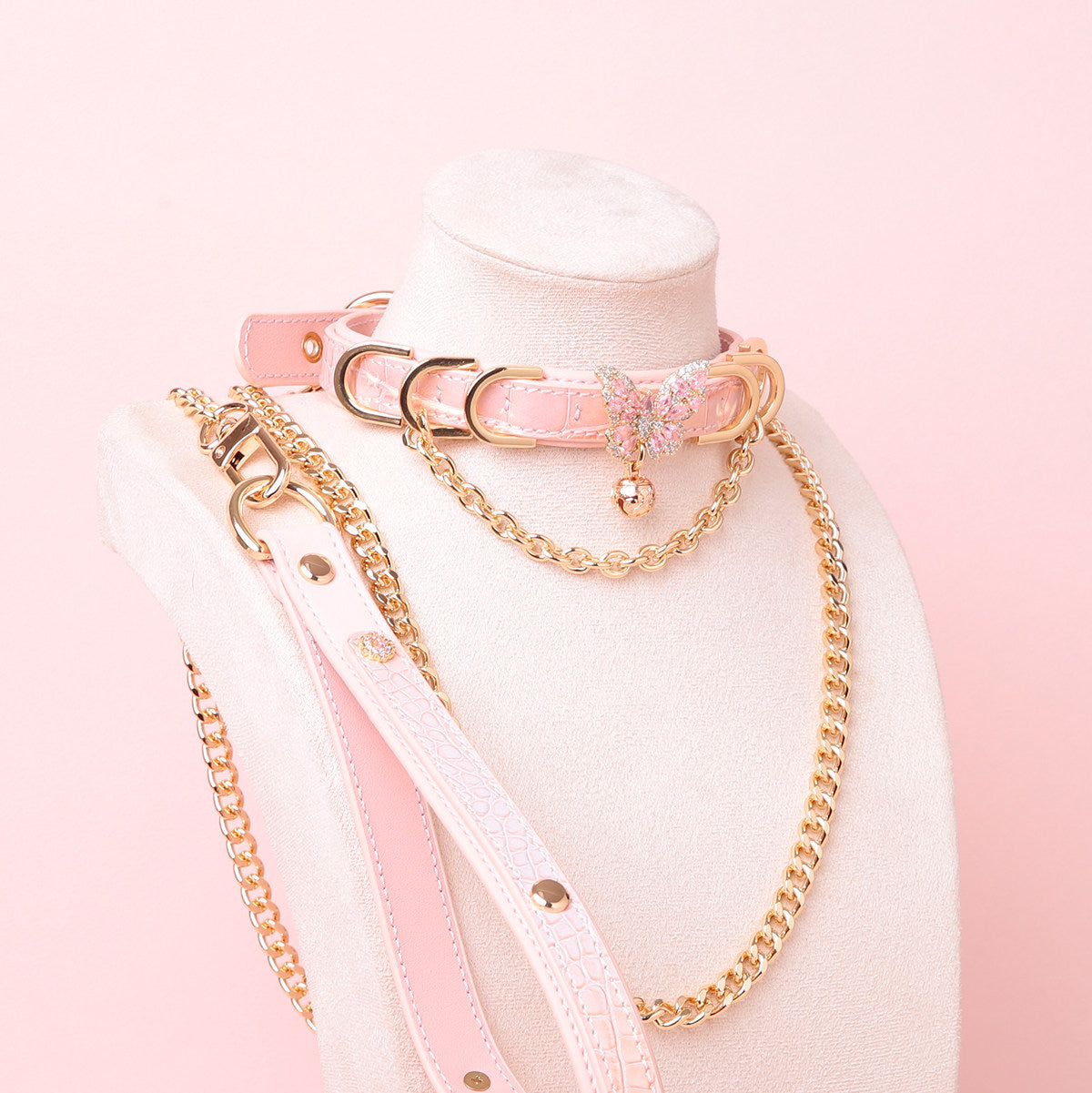 pink and gold leather collar and leash set featuring a gold and pink diamante butterfly charm, gold tone chains, D rings, detachable chain leash and leather hand strap, displayed on a white mannequin against a pink background.