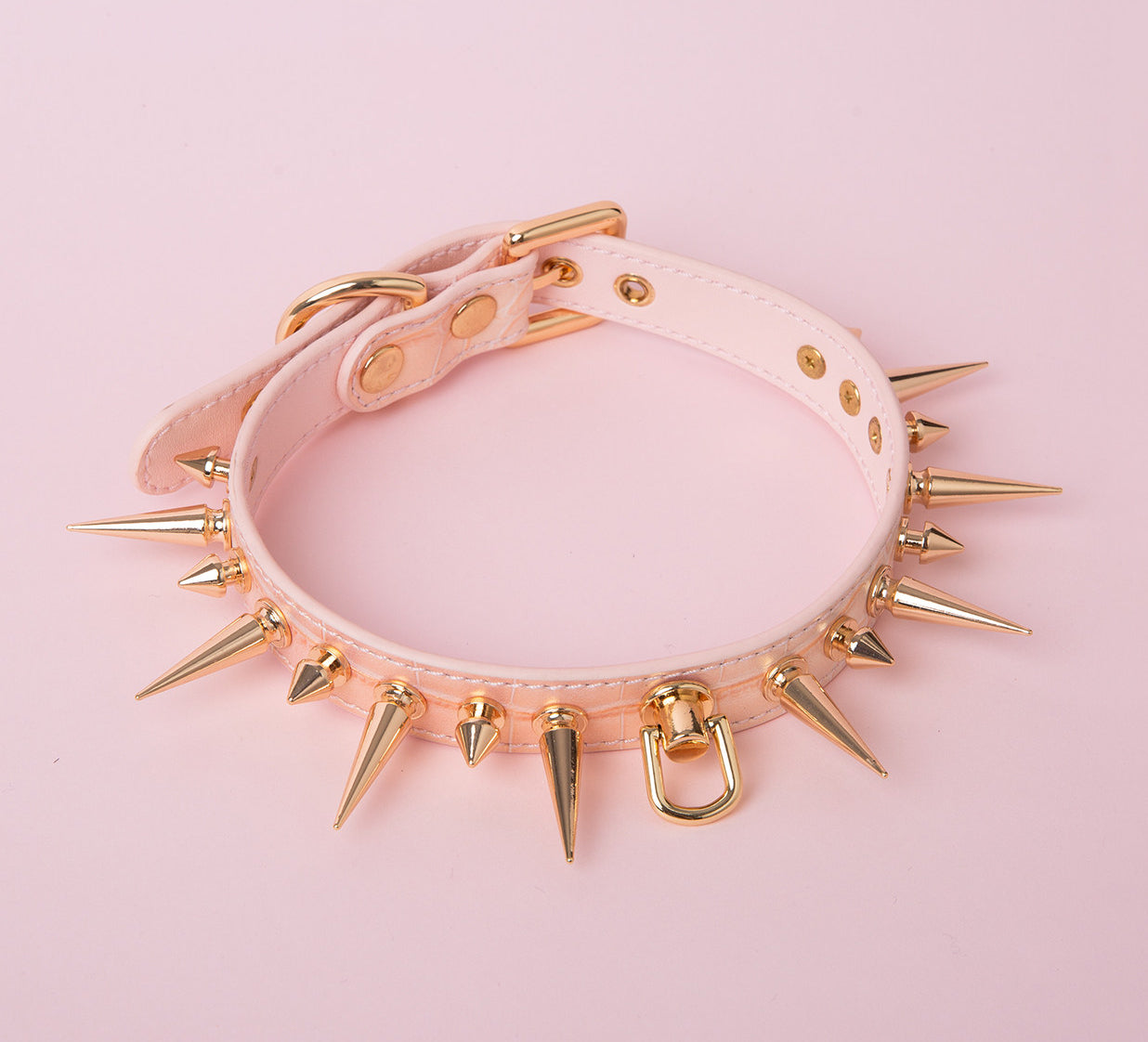 pink and gold leather collar featuring gold-tone alternating long and short spike studs, central D ring attachment and adjustable buckle fastening at the back. displayed against a pink background.