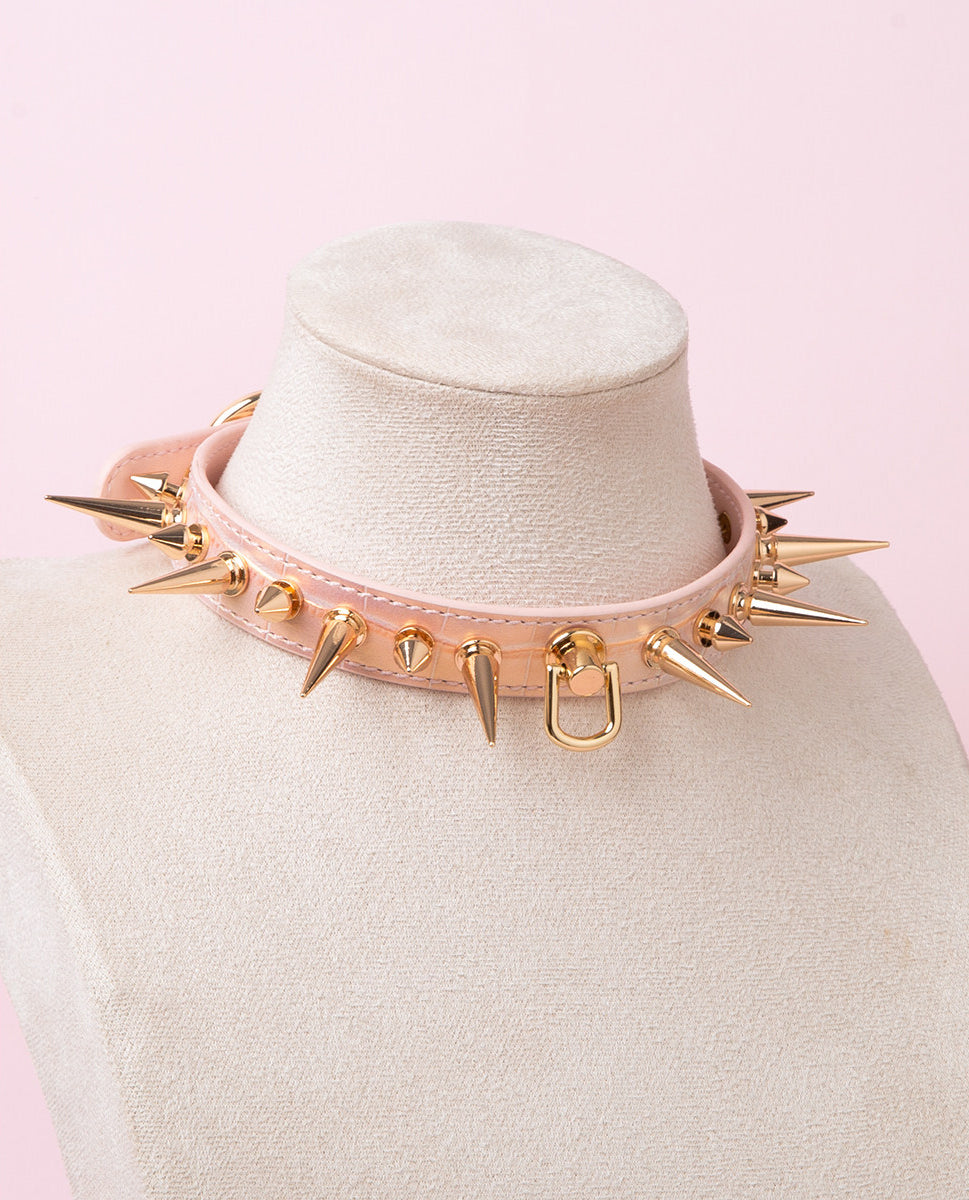pink and gold leather collar featuring gold-tone alternating long and short spike studs and central D ring attachment. displayed on a white mannequin against a pink background.