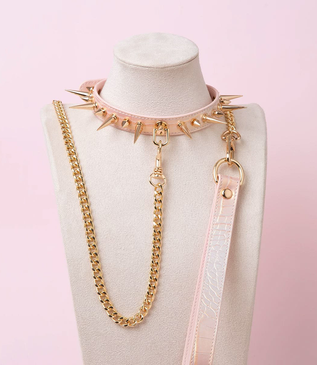 pink and gold leather collar and leash set featuring gold-tone alternating long and short spike studs, D ring attachment with a detachable gold chain leash and pink leather hand strap, displayed on a white mannequin against a pink background.
