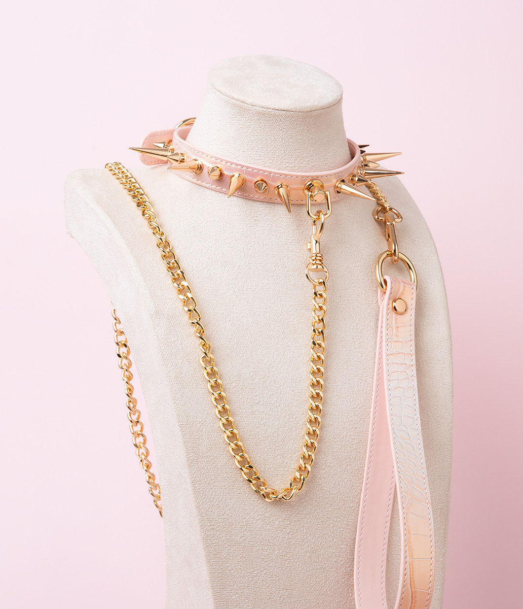 pink and gold leather collar and leash set featuring gold-tone alternating long and short spike studs, D ring attachment with a detachable gold chain leash and pink leather hand strap, displayed on a white mannequin against a pink background.