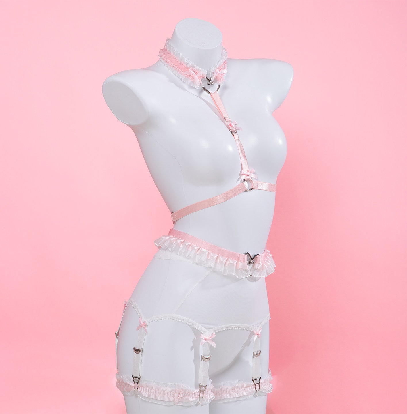 side view of a pink and white harness set featuring ruffled lace, silver heart rings, white mesh suspender belt and satin bow appliques, displayed on a white mannequin against a pink background.