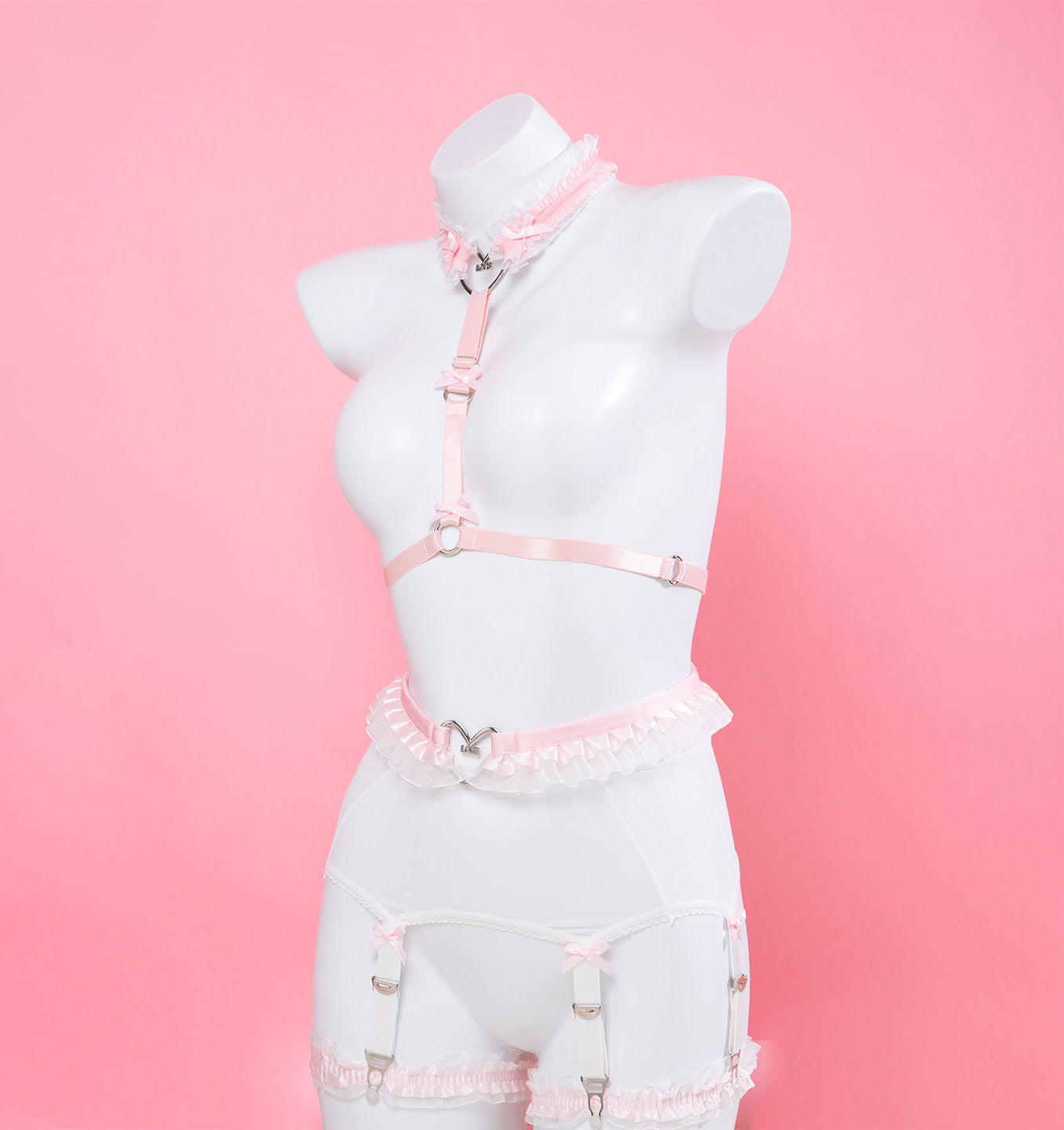 side view of a pink and white harness set featuring ruffled lace, silver heart rings, white mesh suspender belt and satin bow appliques, displayed on a white mannequin against a pink background.
