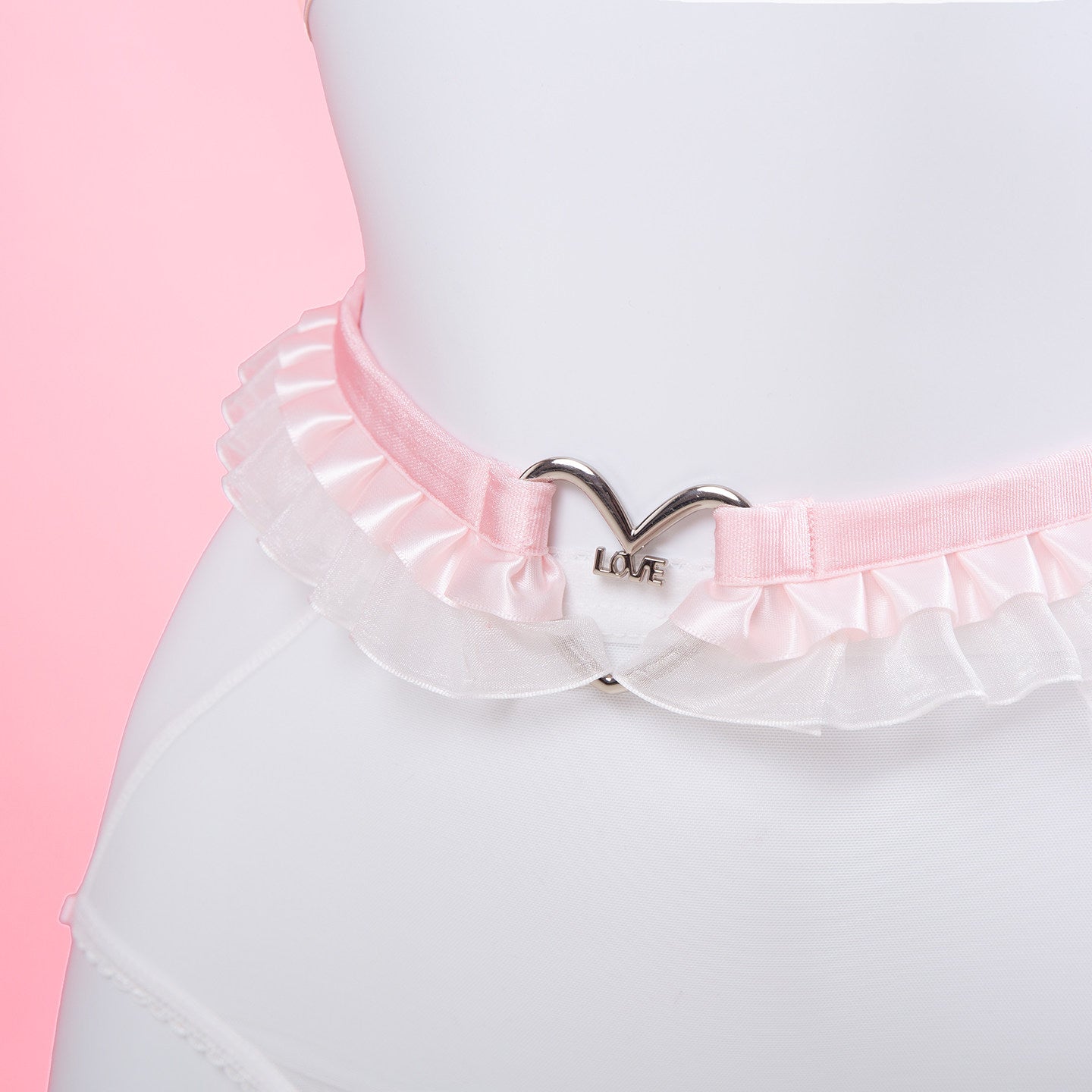 close-up of a white and pink harness set, showing the ruffled lace belt with central silver heart ring hardware, displayed on a white mannequin against a pink background.