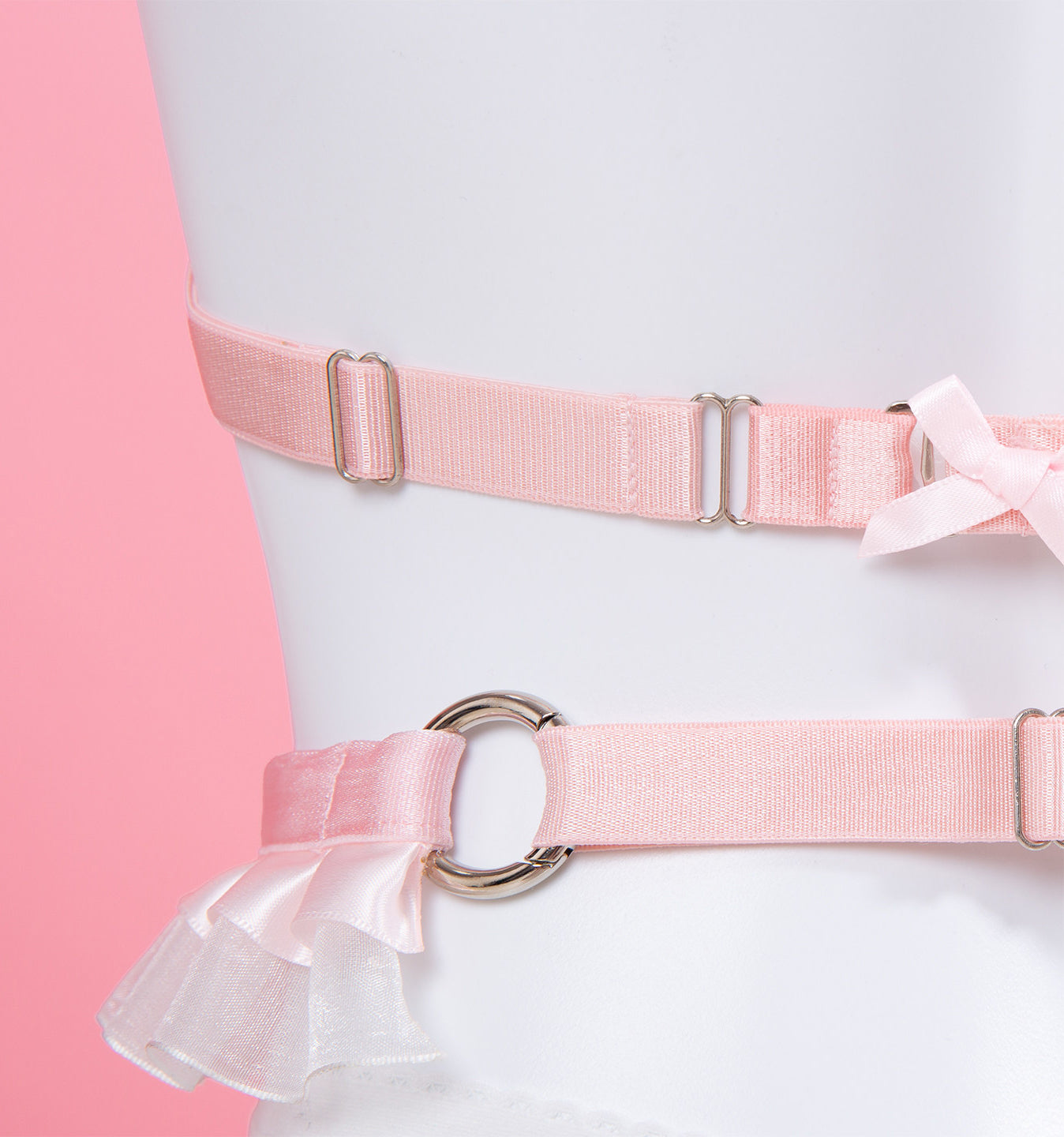 close-up of a white and pink harness set, showing the adjustable pink elastic straps with silver metal sliders, lingerie hook fastening and O ring hardware, displayed on a white mannequin against a pink background.