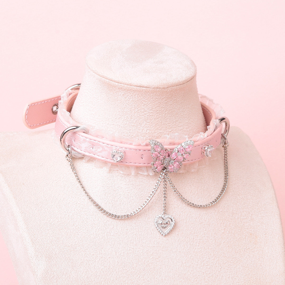 pink leather collar featuring white ruffled lace trim and intricate silver and pink crystal butterfly pendant, D rings, chains and crystal heart charms, displayed on a white mannequin against a pink background.