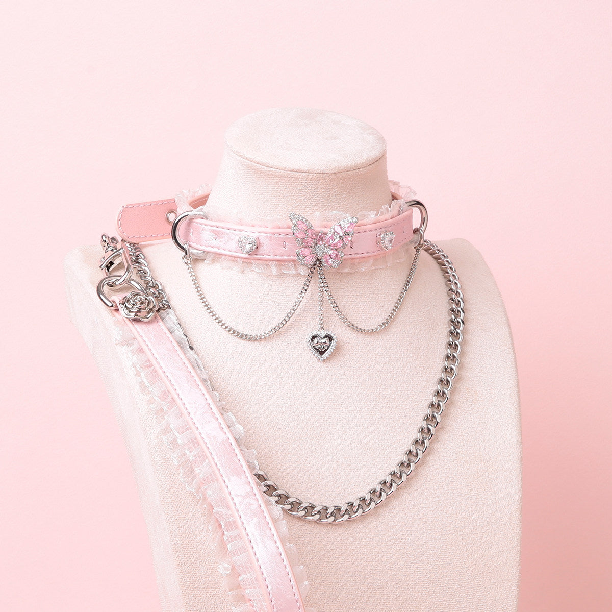 pink leather collar and leash set featuring white ruffled lace trim and intricate silver butterfly and crystal heart charms, with a detachable chain leash and leather hand strap, displayed on a white mannequin against a pink background.
