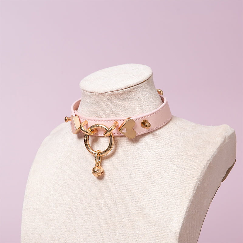 Pink leather collar featuring a central gold tone O ring and detachable gold bell charm. Displayed on a mannequin against a pink background.