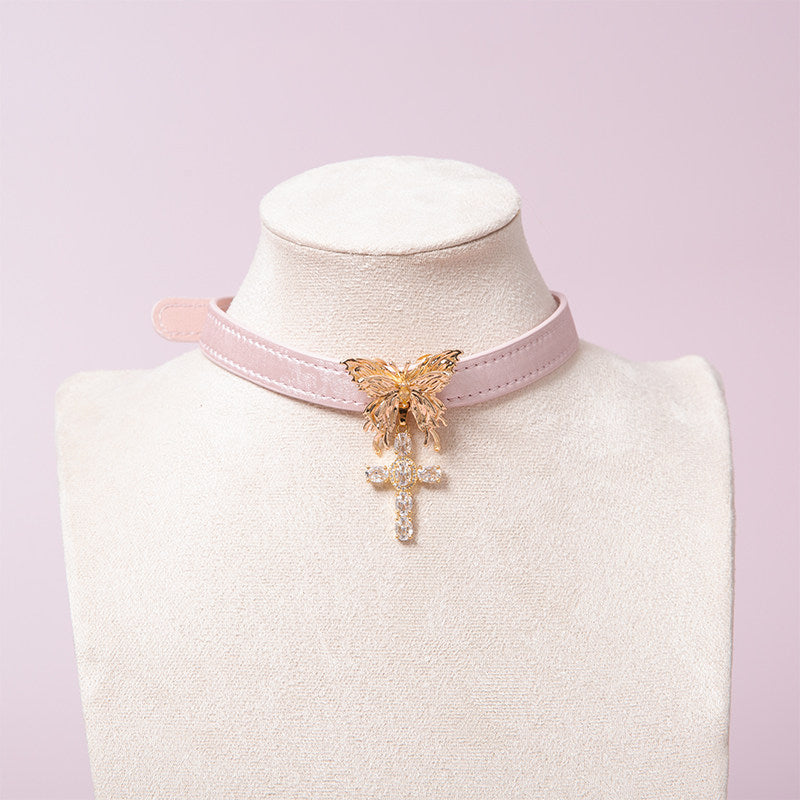 pink and gold collar featuring a gold tone butterfly pendant and gold diamante cross charm, displayed on a white mannequin against a pink background.