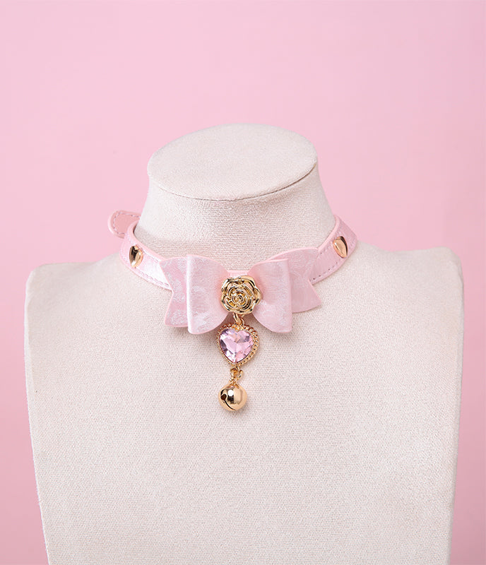 pink and gold collar featuring a ribbon bow shaped centre, gold rose and heart studs with a pink and gold crystal heart and bell charm, displayed on a white mannequin against a pink background.