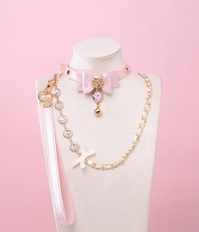 pink and gold collar and leash set featuring a ribbon bow shaped centre, gold rose and heart studs, pink and gold crystal heart and bell charm with a detachable chain and crystal leash and leather hand strap, displayed on a white mannequin against a pink background.