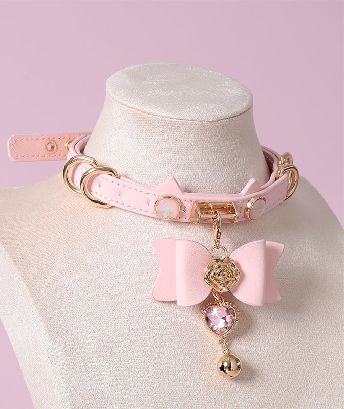 Pink and gold cat girl collar featuring a unique cat ear shaped choker, gold tone hardware and detachable pink and gold bow pendant with pink crystal heart charm. Displayed on a mannequin against a pink background.