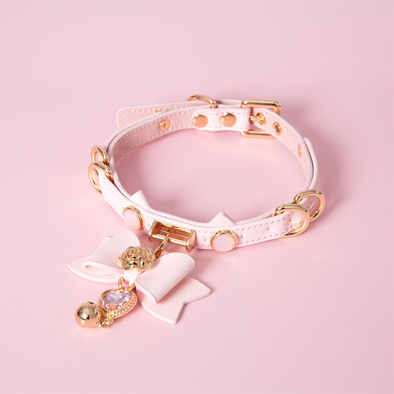 Pink and gold cat girl collar featuring a unique cat ear shaped choker, gold tone hardware and detachable pink and gold bow pendant with pink crystal heart charm. Displayed on a pink background.