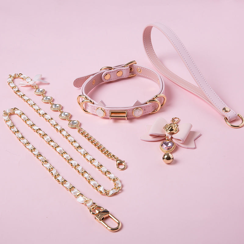 Pink and gold cat girl collar and leash set featuring a unique cat ear shaped choker, detachable pink and gold bow, heart and bell pendant, detachable crystal and chain leash and pink leather hand strap. Displayed on a pink background.