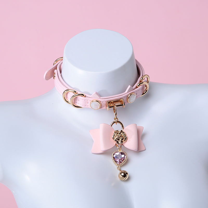 Pink and gold cat girl collar featuring a unique cat ear shaped choker, gold tone hardware and detachable pink and gold bow, heart and bell pendant. Displayed on a mannequin against a pink background.