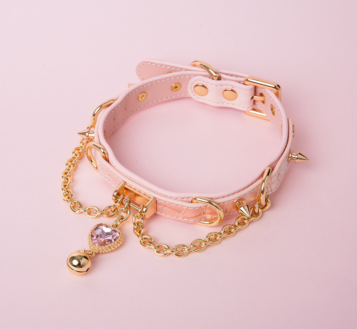 pink and gold leather collar featuring a pink and gold crystal heart charm, gold bell, chains and spike studs, displayed against a pink background.