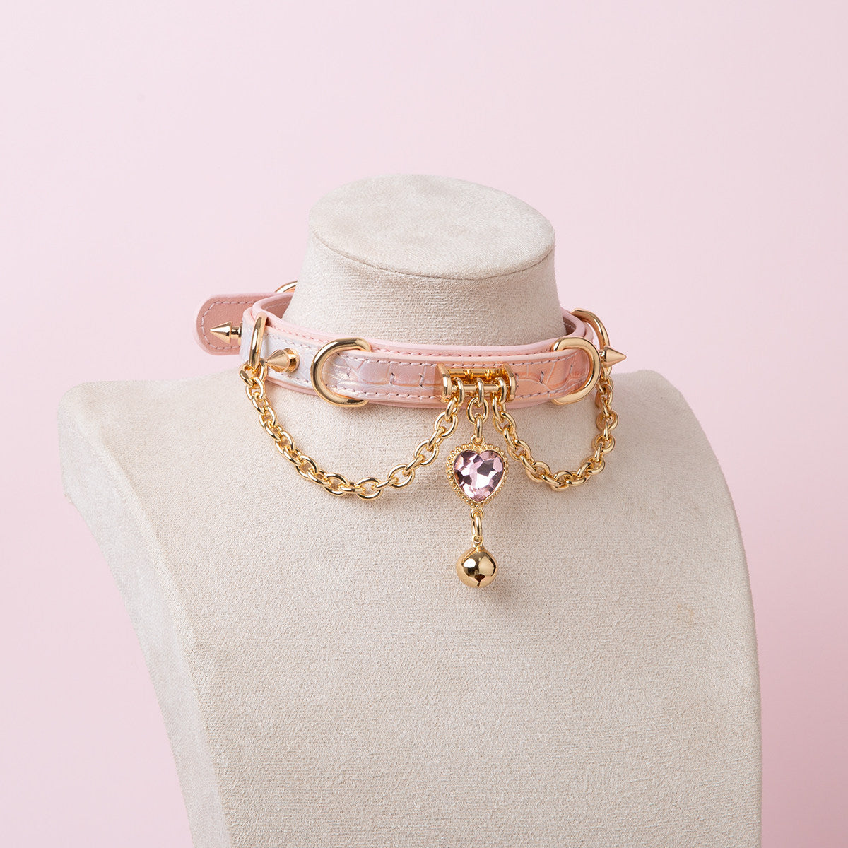 pink and gold leather collar featuring a pink and gold crystal heart charm, gold bell, chains and spike studs, displayed on a white mannequin bust against a pink background.
