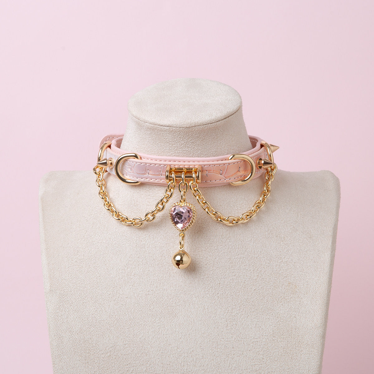pink and gold leather collar featuring a pink and gold crystal heart charm, gold bell, chains and spike studs, displayed on a white mannequin bust against a pink background.
