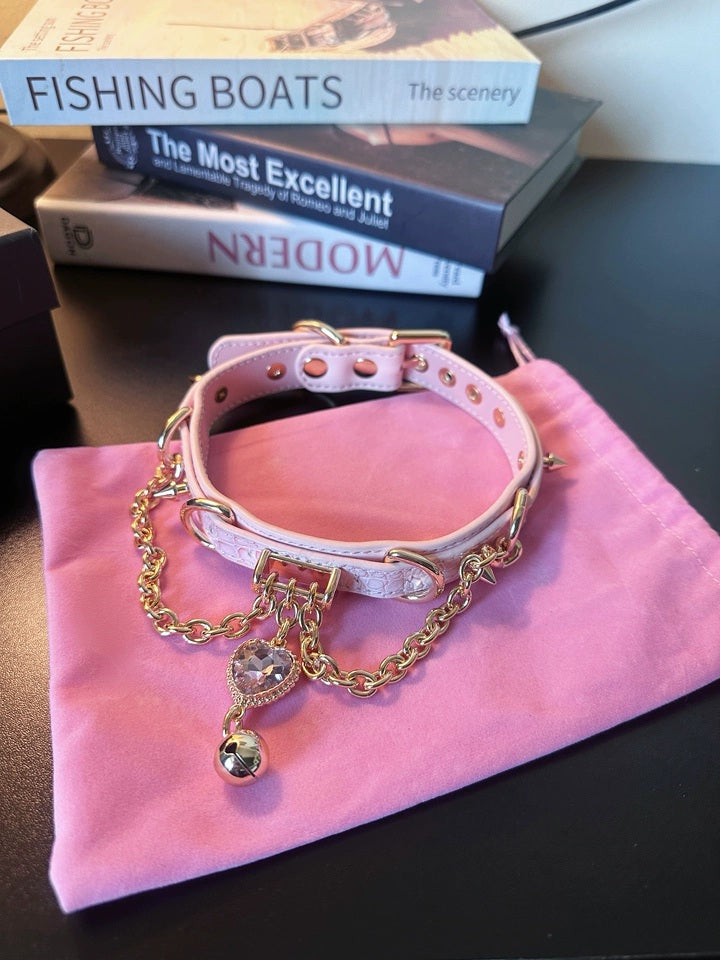 pink and gold leather collar featuring a crystal heart charm, gold bell, chains and spike studs, displayed on top of a pink velvet drawstring storage bag.