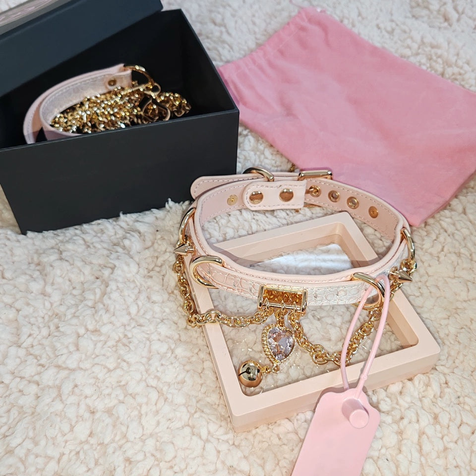 pink and gold leather collar featuring a crystal heart charm, gold bell, chains and spike studs, displayed on top of a pink velvet drawstring bag and storage box.