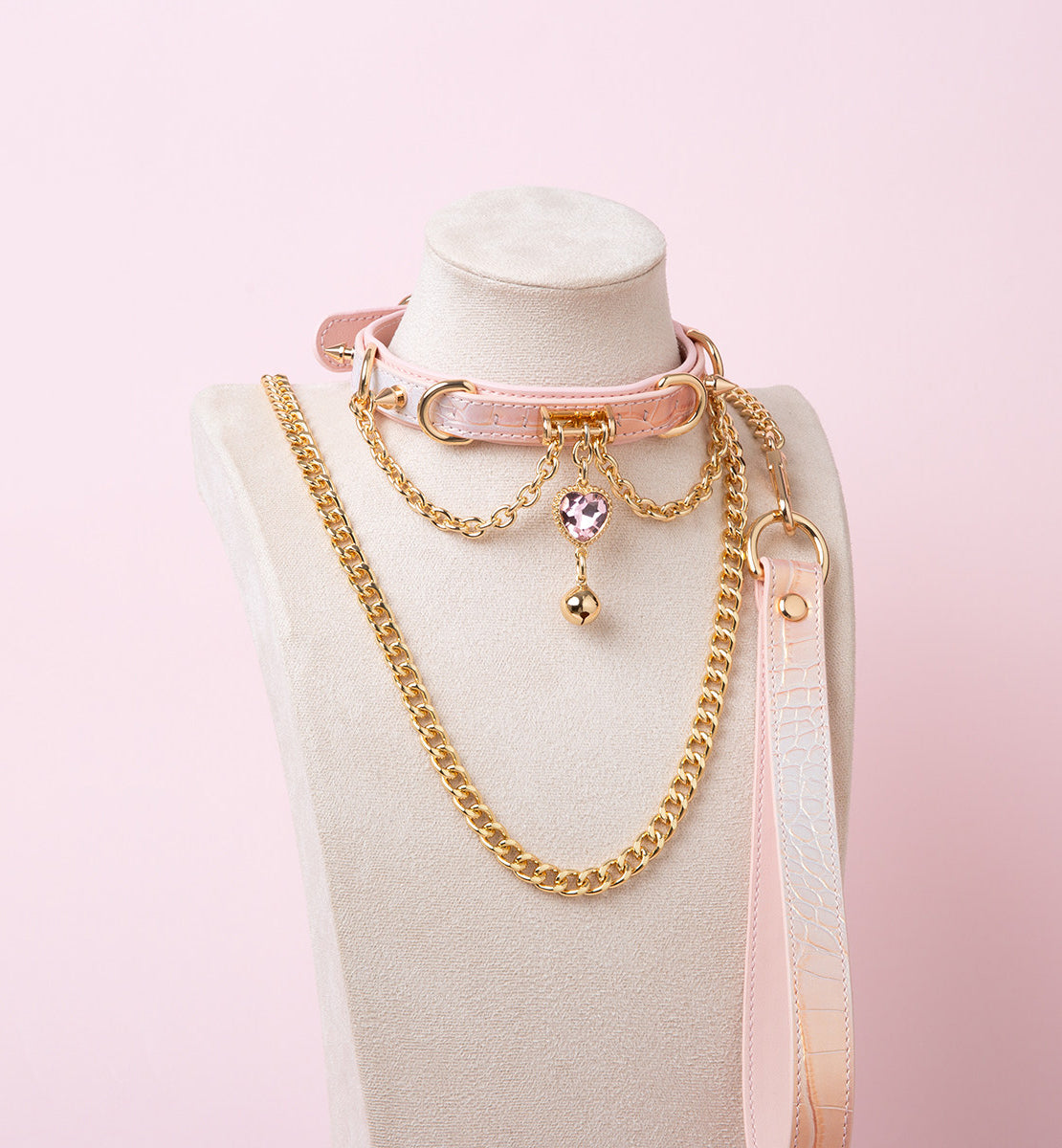pink and gold collar and leash set featuring a pink and gold crystal heart charm, gold bell, chains and pink leather hand strap, displayed on a white mannequin bust against a pink background.
