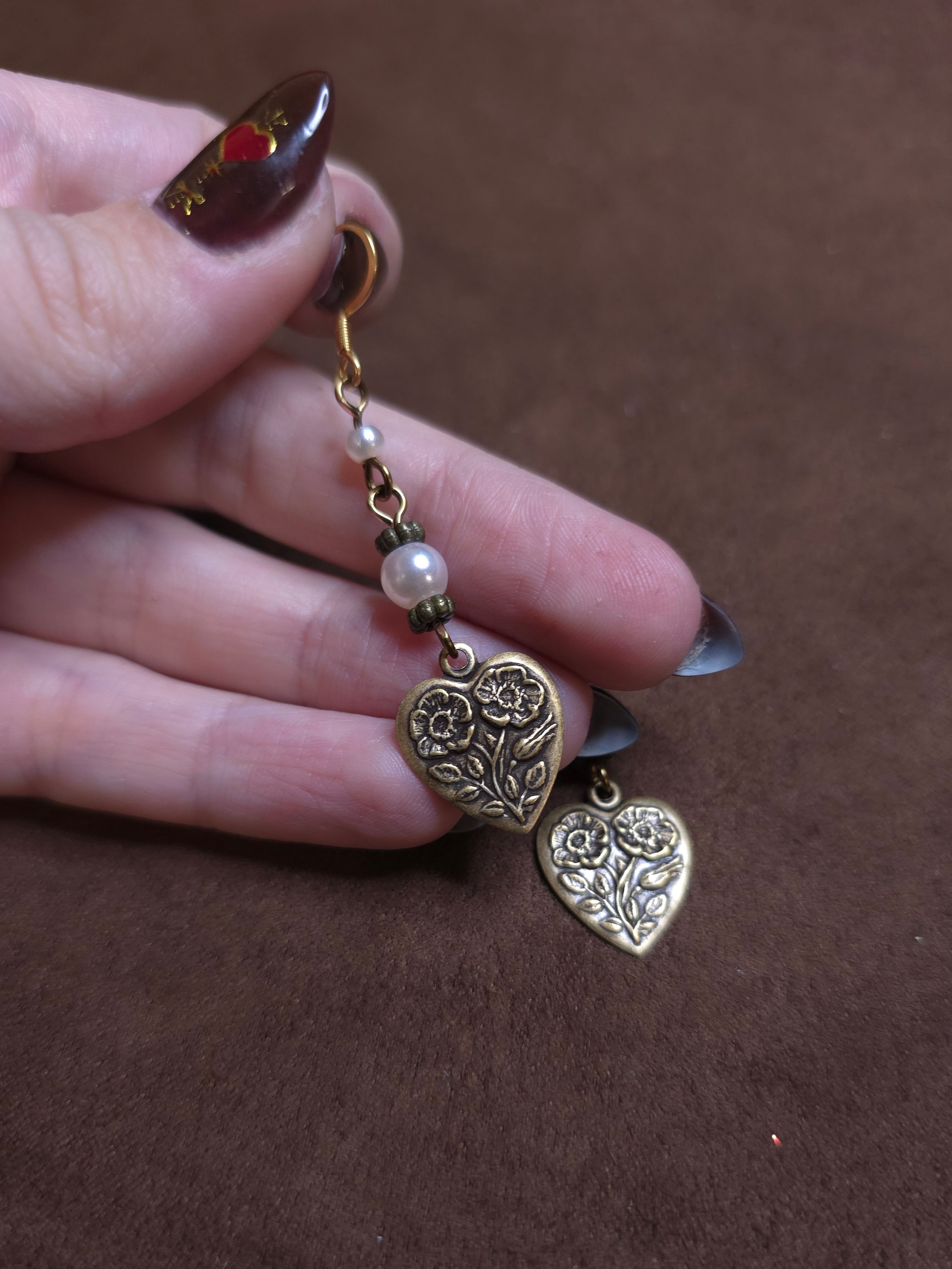 Hand holding a pair of heart-shaped earrings with intricate designs on a brown background