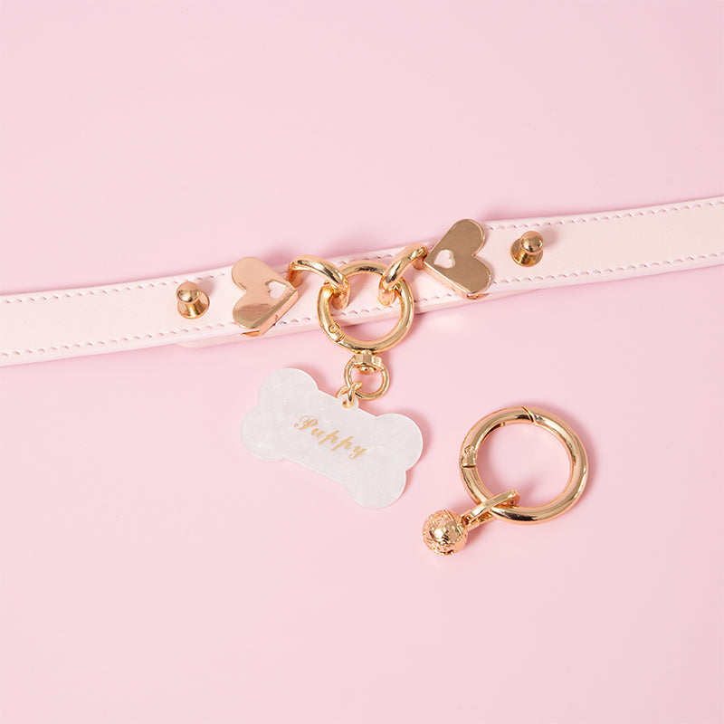 Pink leather collar featuring a central gold tone O ring, detachable custom engraved bone shaped pet tag and gold bell charm. Displayed against a pink background.